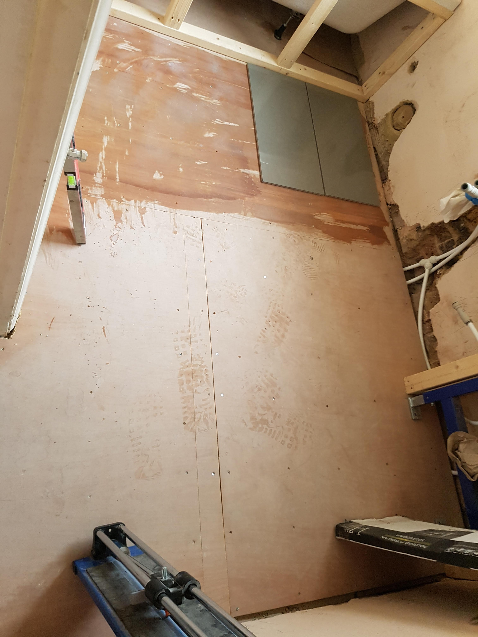 Przygotowanie ścian łazienki pod płytki: płyta OSB, częściowo położone płytki, widoczne rury instalacyjne i poziomica przy ścianie, ślady butów na podłodze, narzędzie do cięcia płytek.