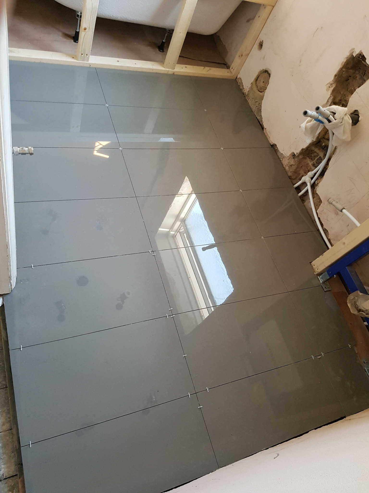 Ułożone szare płytki podłogowe w trakcie prac wykończeniowych w łazience, widoczne krzyżyki dystansowe, instalacja wodna w ścianie i fragment wanny.