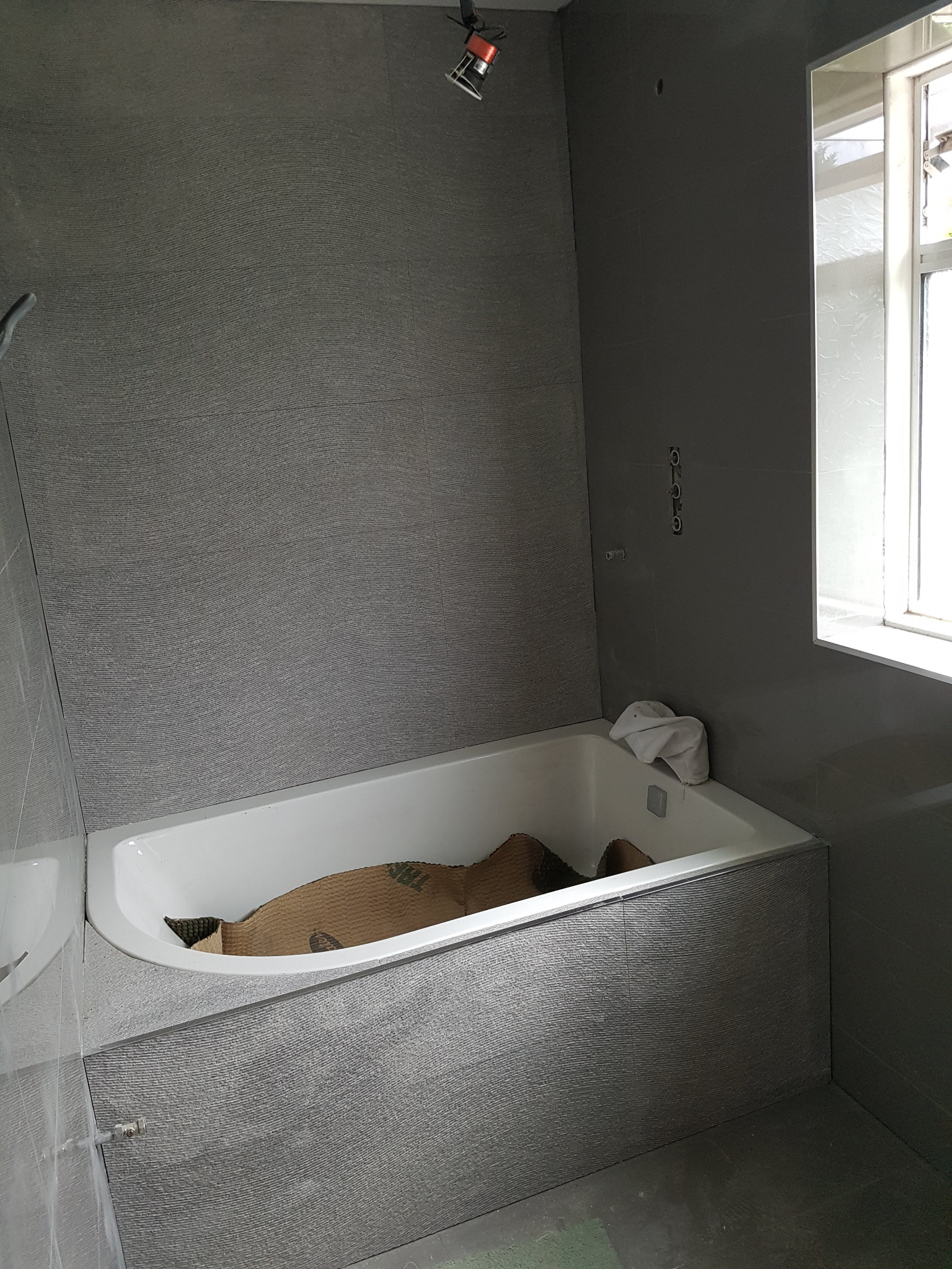 Szara łazienka w trakcie remontu z wanną zabezpieczoną kartonem, widoczne płytki na ścianach, biała wanna i okno.