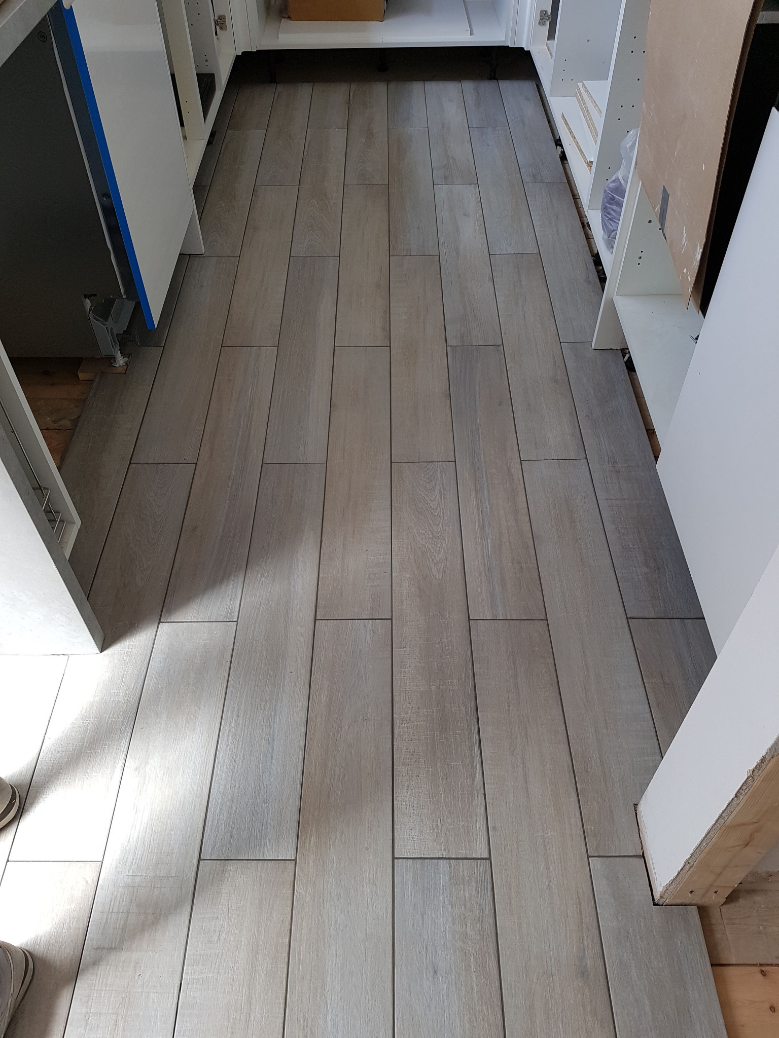 Ułożona podłoga z płytek imitujących drewno w wąskim pomieszczeniu, widoczne fragmenty białych szafek kuchennych, w tle kartonowe pudło.