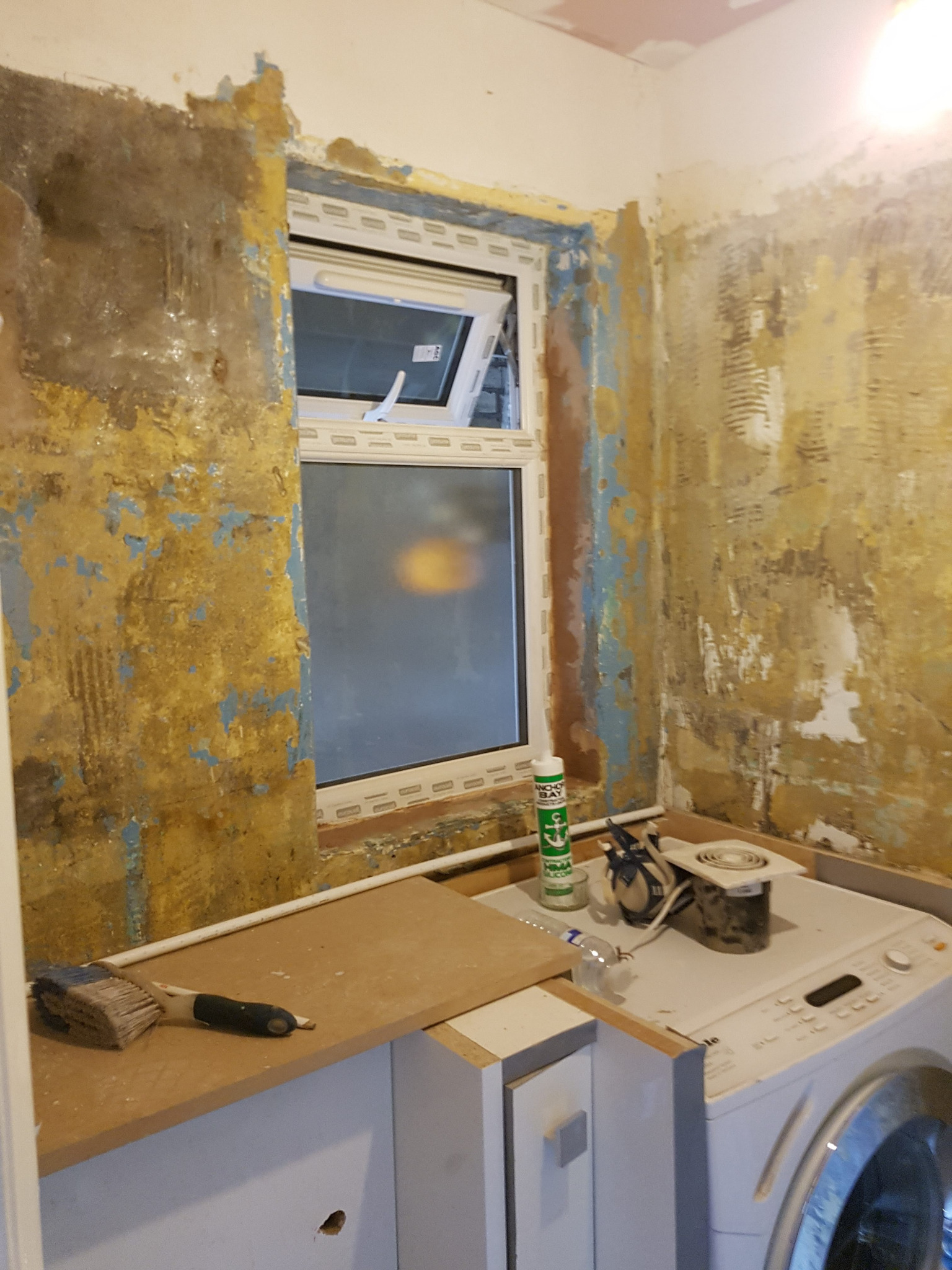 Ściana z resztkami starej farby, okno PCV uchylone, pralka, pędzel i tuba silikonu w trakcie prac przygotowawczych do remontu.