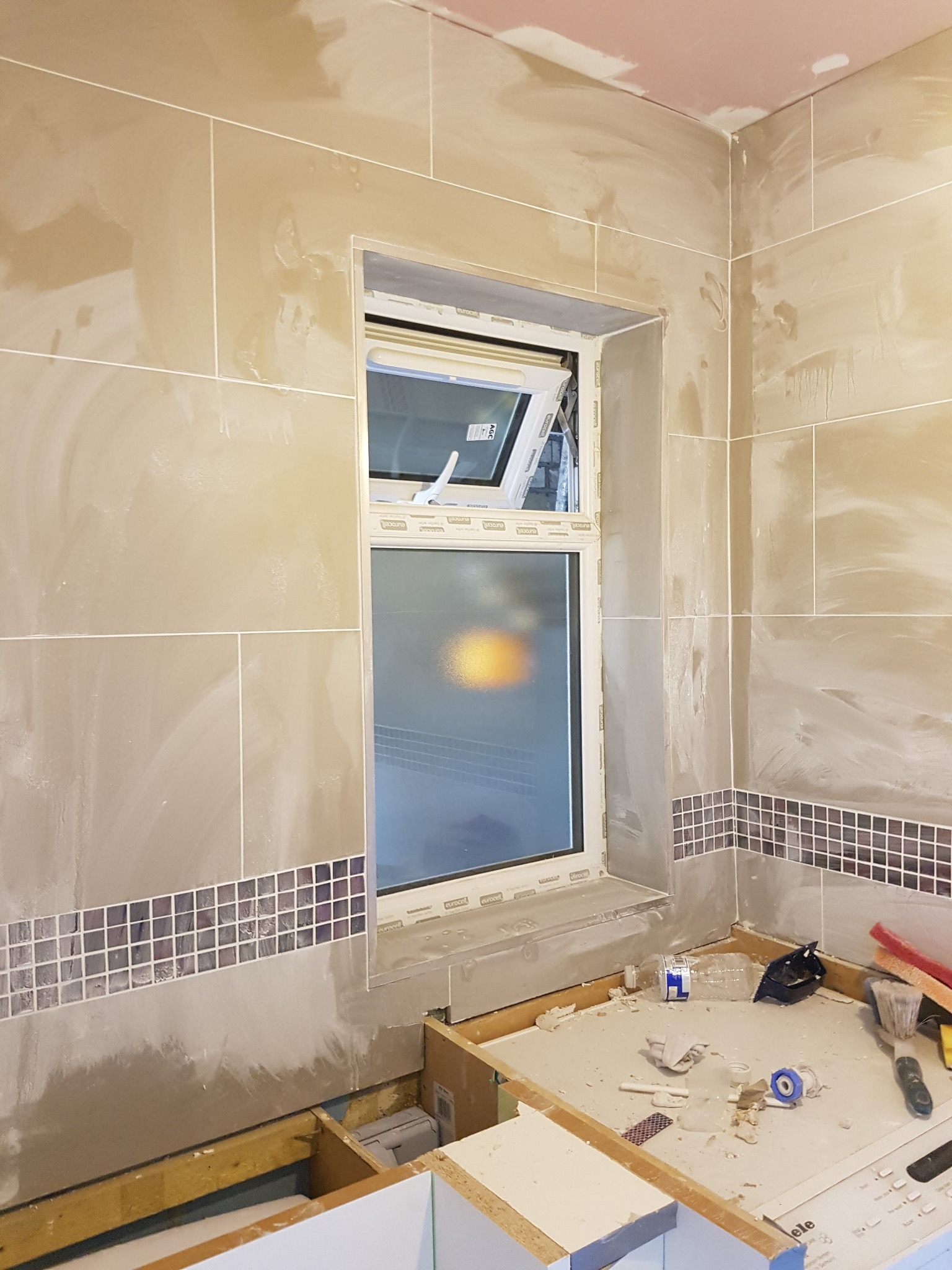 Ściana łazienki w trakcie prac wykończeniowych, widoczne beżowe płytki, mozaika, otwarte okno i narzędzia na blacie.