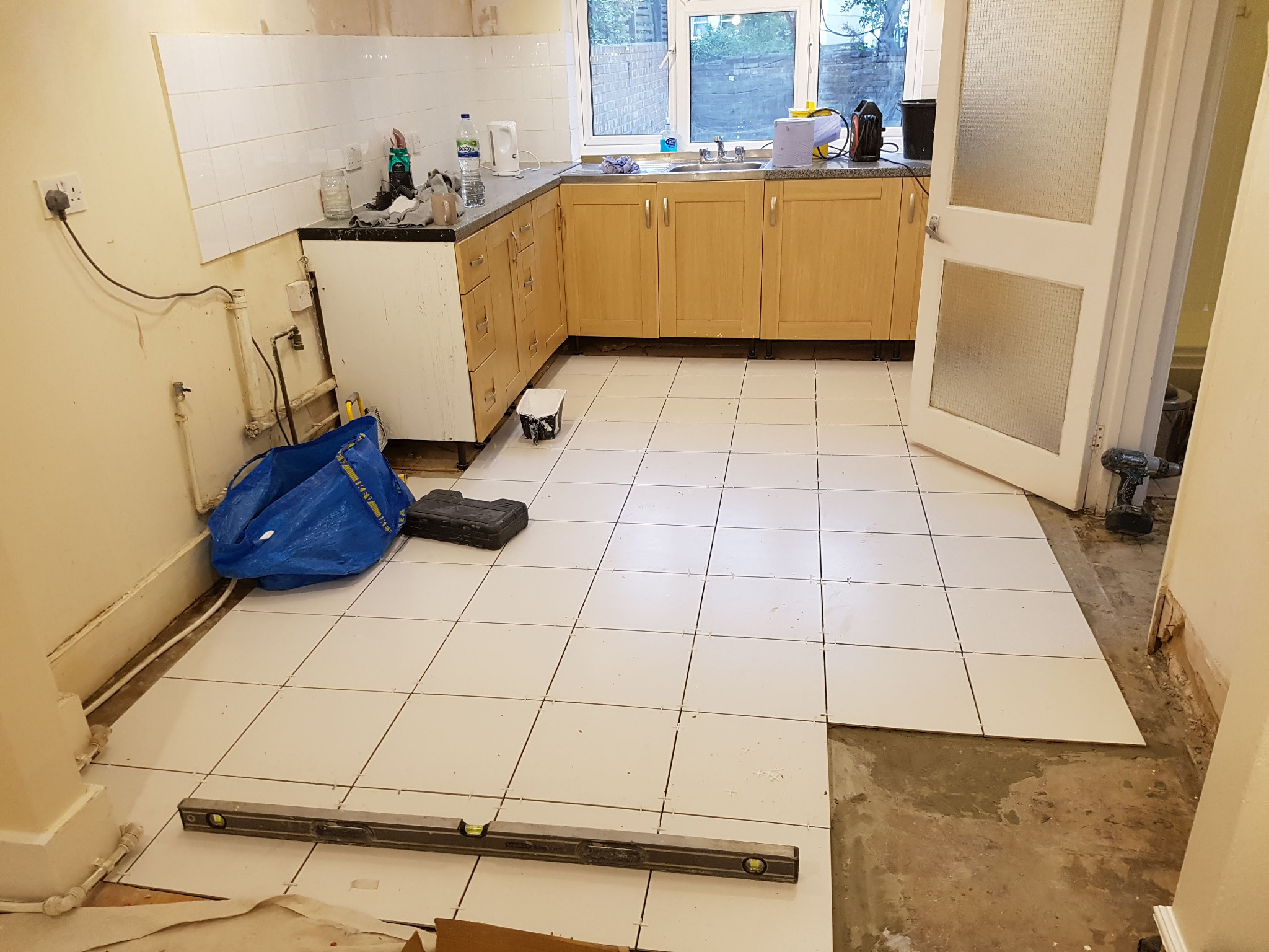 Układanie białych płytek na podłodze w kuchni podczas remontu, widoczny poziomica, torba budowlana i narzędzia