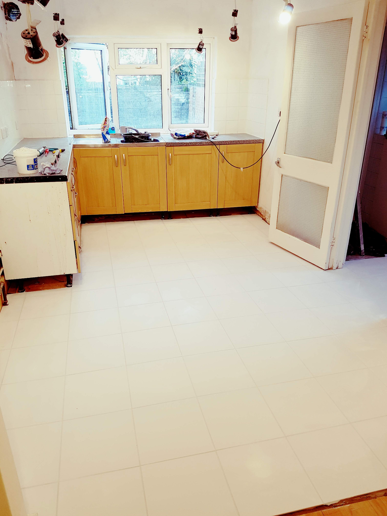 Jasna kuchnia w trakcie prac remontowych: świeżo położone białe płytki podłogowe, drewniane szafki, okno z widokiem na zieleń, oraz widoczne przygotowania do montażu oświetlenia sufitowego.