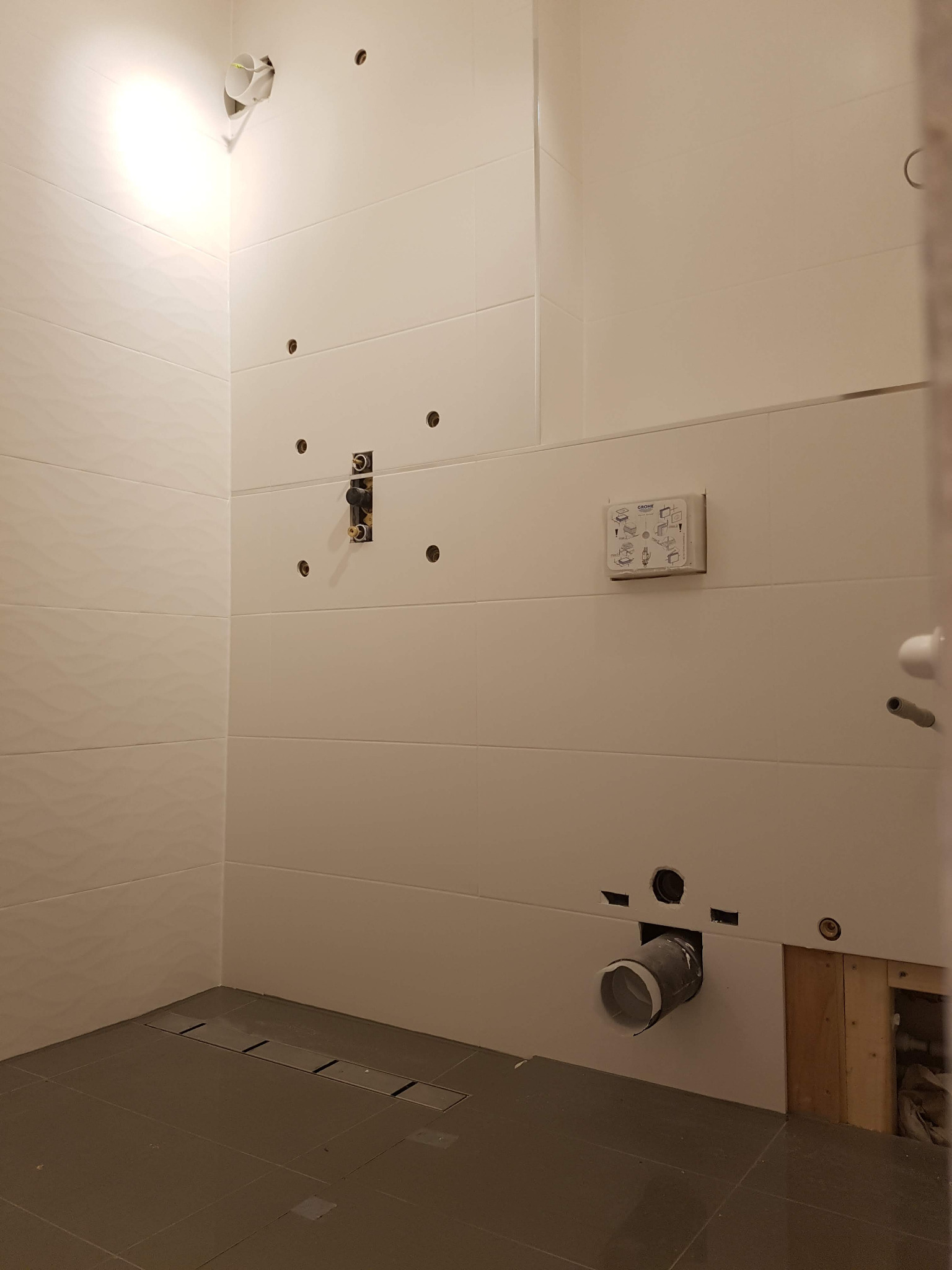 Wnętrze łazienki w trakcie prac: białe płytki z delikatnym wzorem, szara podłoga z odpływem liniowym, widoczne instalacje wodno-kanalizacyjne, otwory w ścianach, instrukcja montażu armatury Grohe.