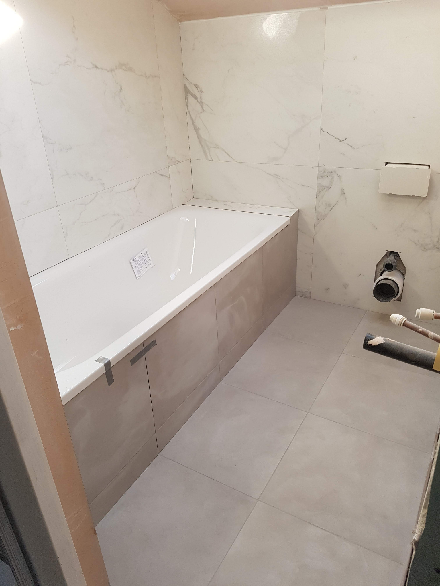 Wanna obudowana płytami, z widocznymi taśmami maskującymi, zamontowana w łazience wyłożonej marmurowymi płytkami, z widocznymi elementami instalacji wodno-kanalizacyjnej, podczas prac wykończeniowych.