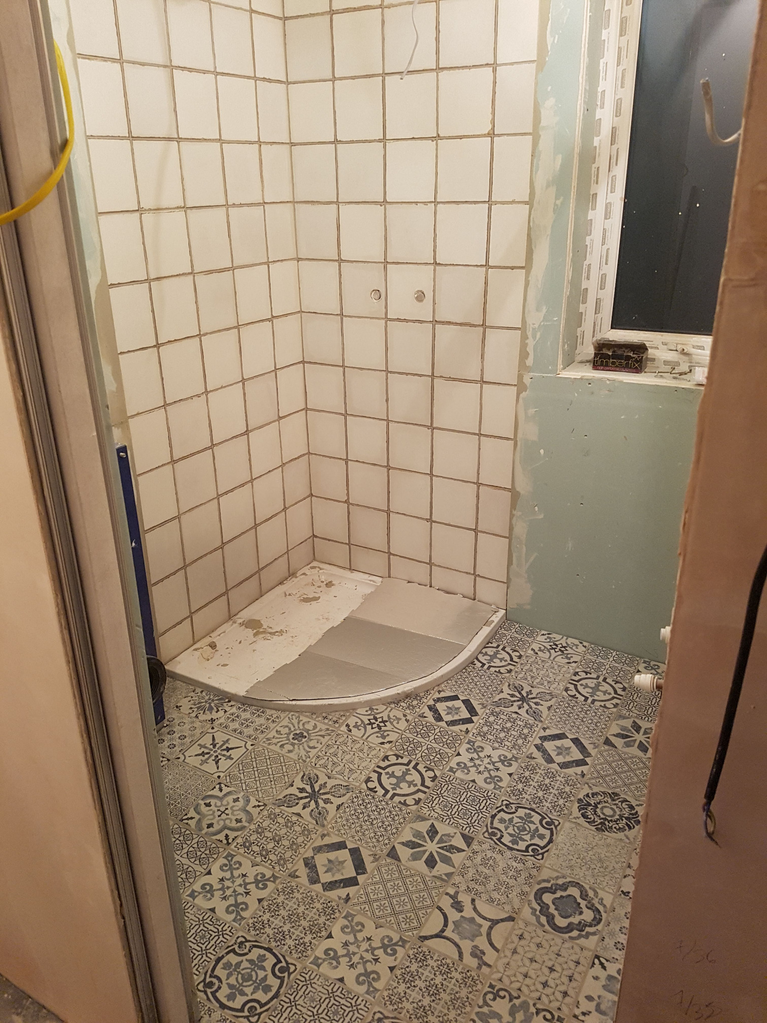 Wnętrze łazienki w trakcie prac: białe płytki ścienne w kabinie prysznicowej, wzorzysta podłoga w odcieniach szarości i błękitu, widoczne ślady zaprawy i niedokończone wykończenia ścian.