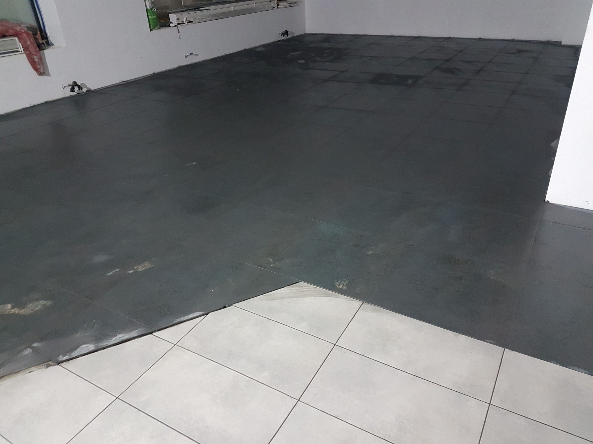 Częściowo ułożona podłoga z ciemnych płytek w pomieszczeniu, widoczne fragmenty białych płytek, ślady po kleju i fugowaniu.