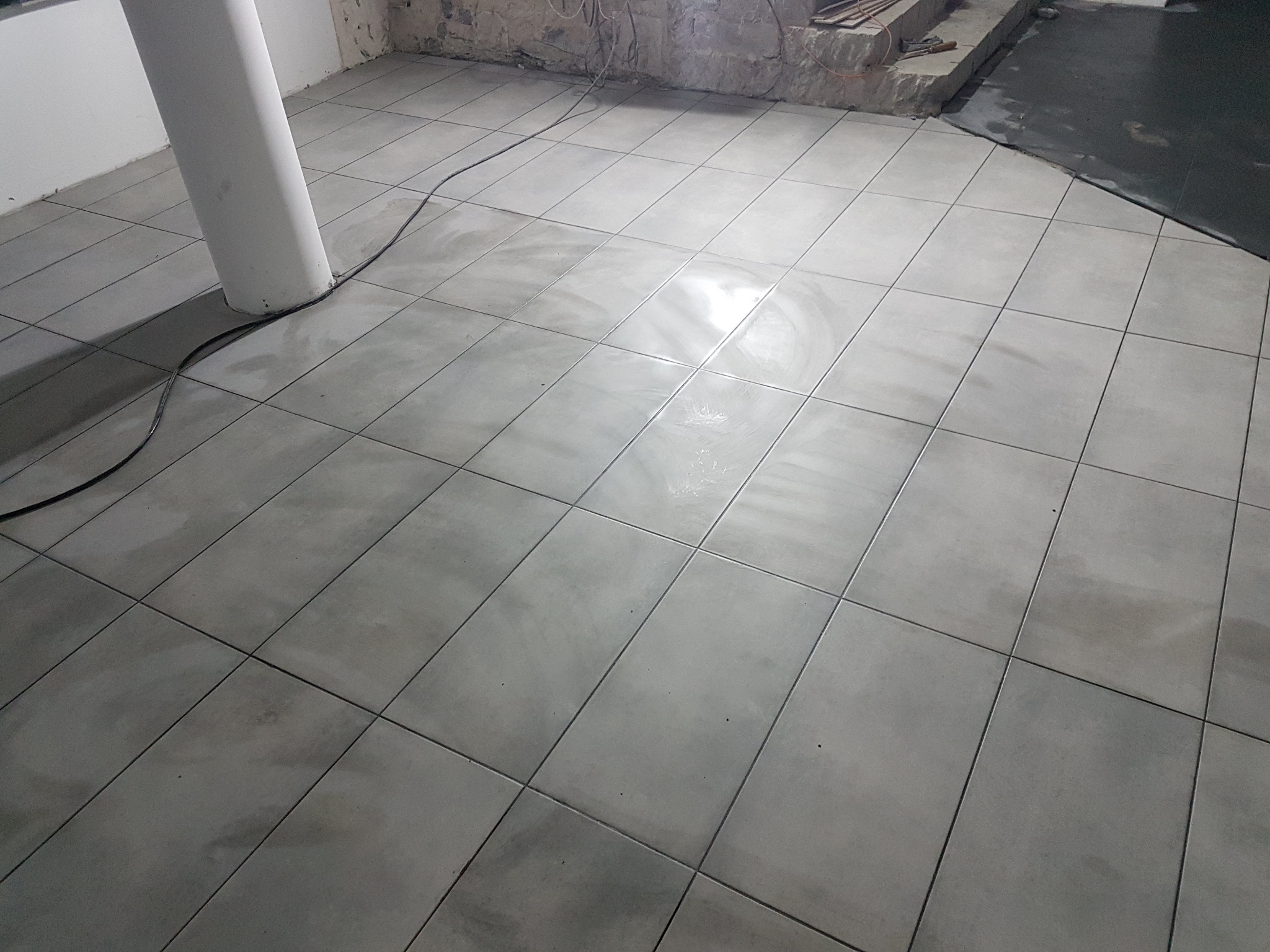 Ułożone szare płytki podłogowe w pomieszczeniu z kolumną i surową ścianą w tle, widoczne kable.
