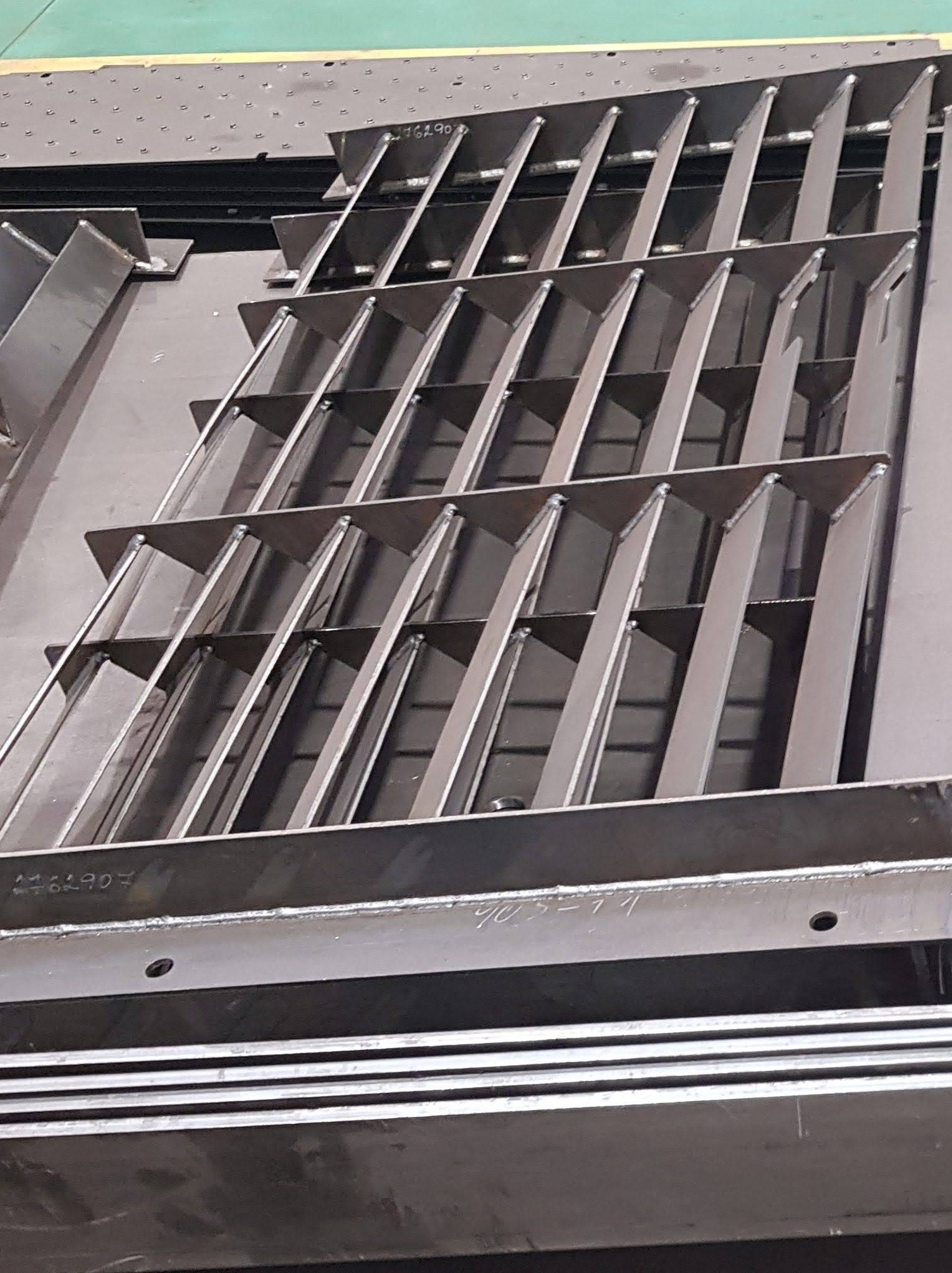 Metalowa konstrukcja kratowa z widocznymi spawami i numerami seryjnymi naniesionymi na elementach, leżąca na metalowej powierzchni w warsztacie.