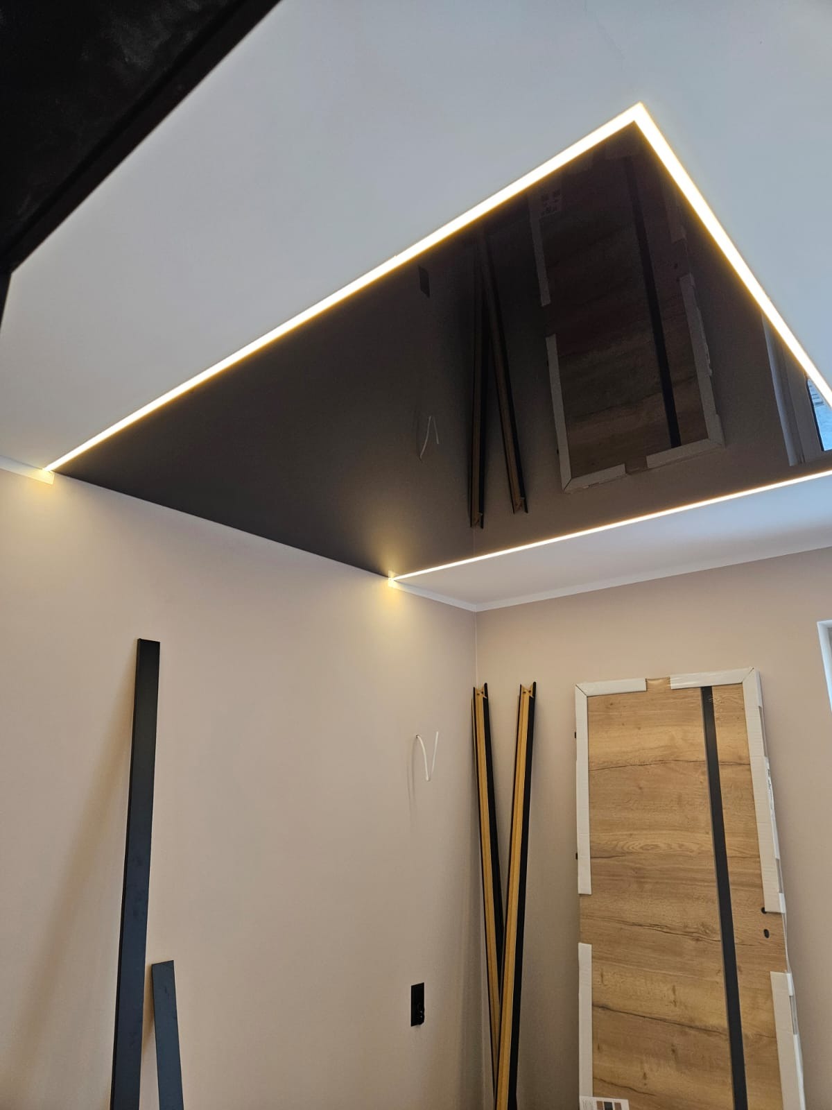 Błyszczący, czarny sufit napinany z oświetleniem LED w kształcie prostokąta, odbijający elementy pokoju. Widoczne listwy przypodłogowe i drzwi.