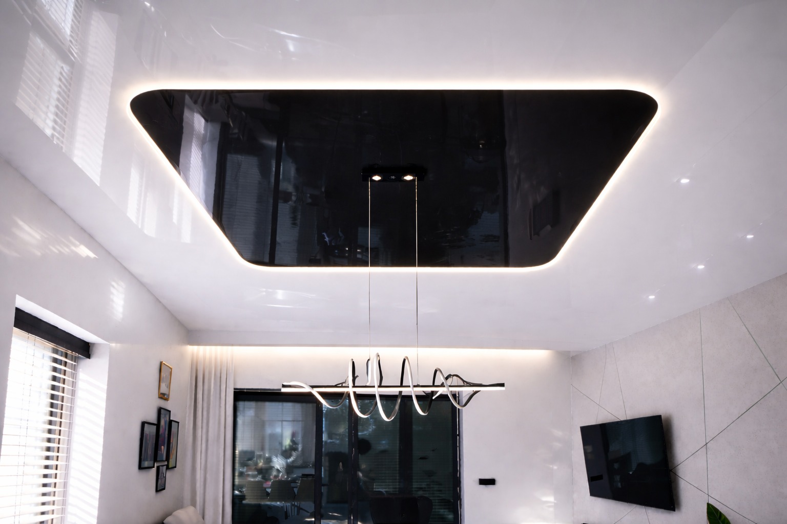 Biały sufit napinany z czarną, podświetloną wnęką w kształcie trapezu i nowoczesną lampą wiszącą. Widok na okno i ścianę z telewizorem.