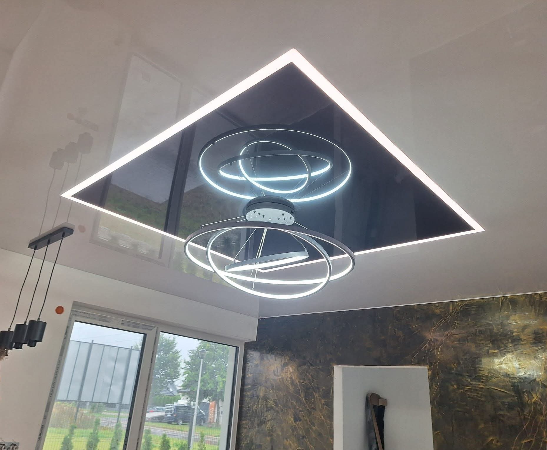 Biały, błyszczący sufit napinany z czarnym wkładem i oświetleniem LED w kształcie kwadratu. Dodatkowo nowoczesna lampa wisząca z okręgami LED. Widok z dołu.