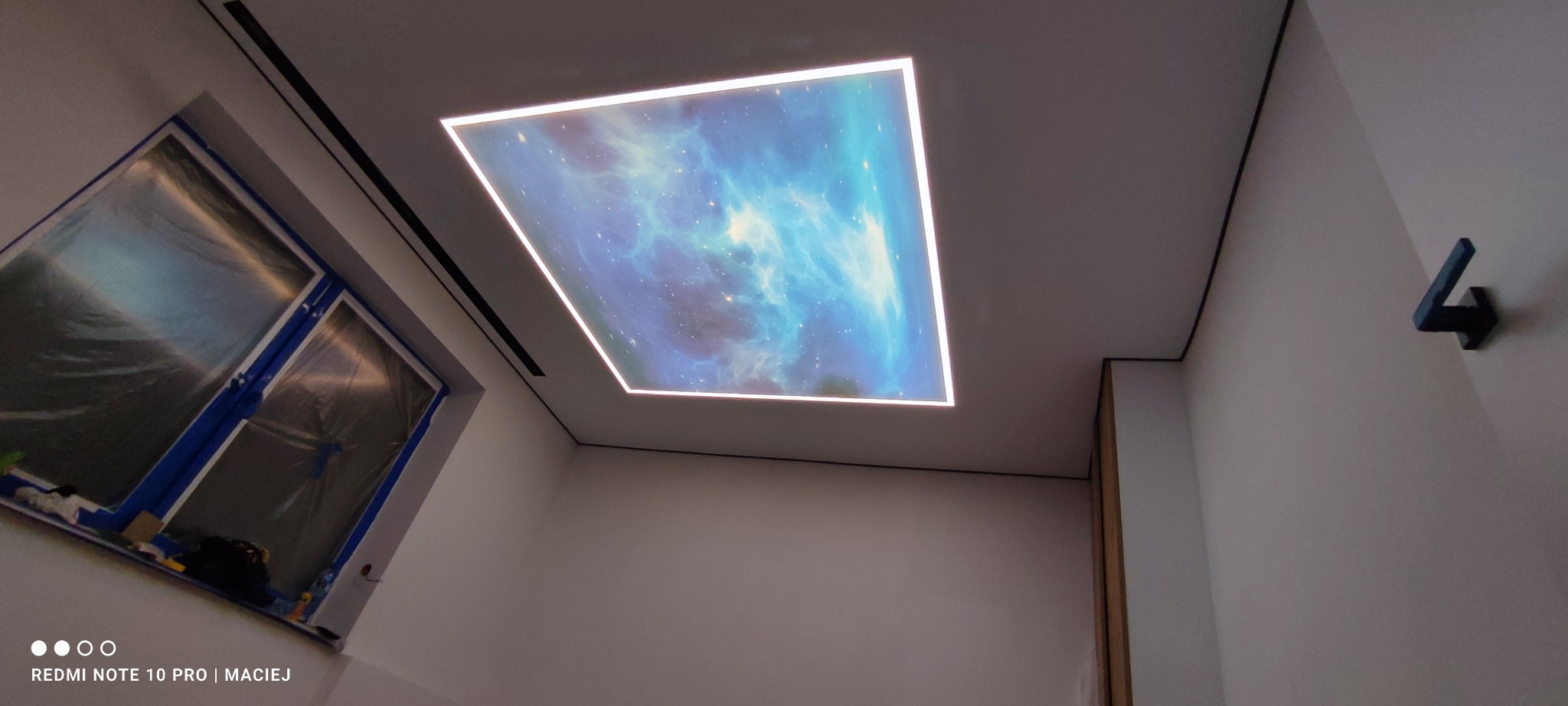 Podświetlany sufit napinany z motywem kosmosu w nowoczesnym wnętrzu. Okno zabezpieczone folią malarską, minimalistyczny wieszak na ścianie. Ujęcie z dołu.