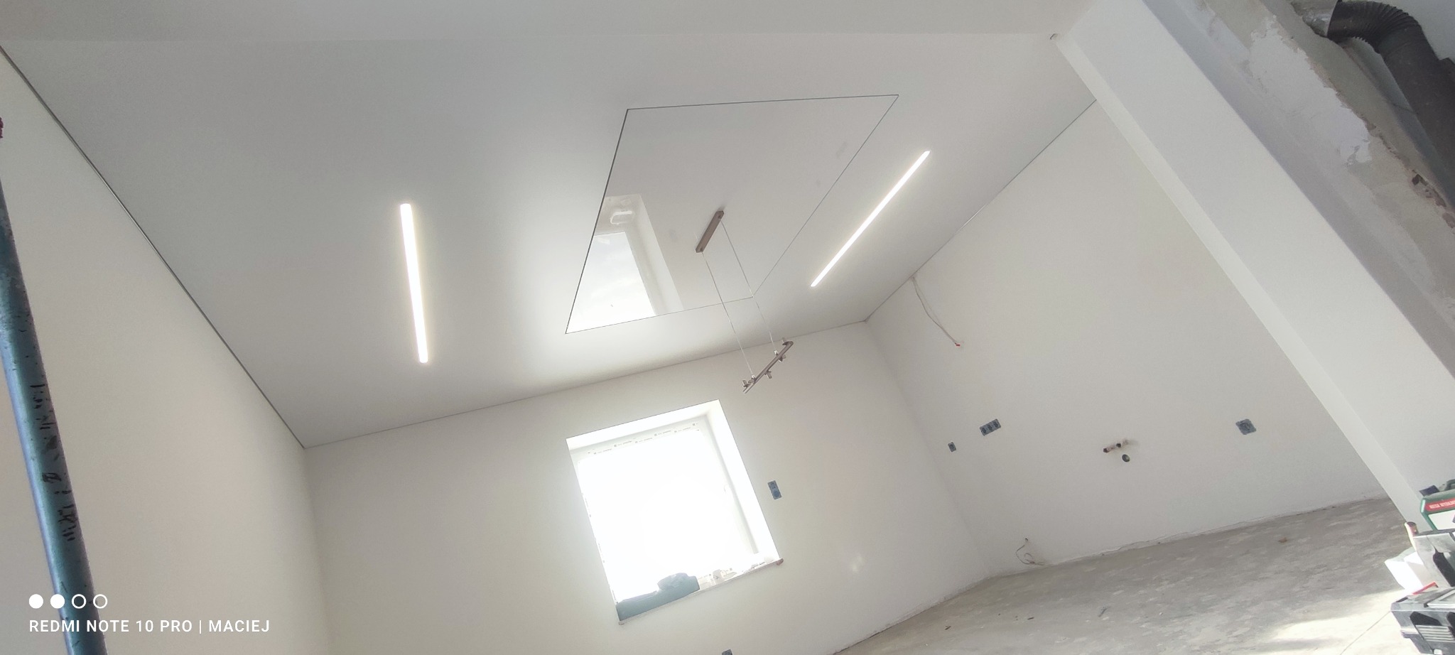 Biały sufit z oświetleniem LED i otworem rewizyjnym. Widoczne okno i fragment ściany z gniazdkami. Ujęcie z dołu, podkreślające nowoczesny design.