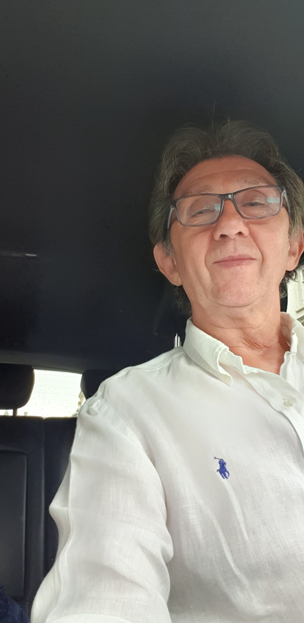 Portret mężczyzny w okularach i białej koszuli z logo, siedzącego w samochodzie z czarną tapicerką.