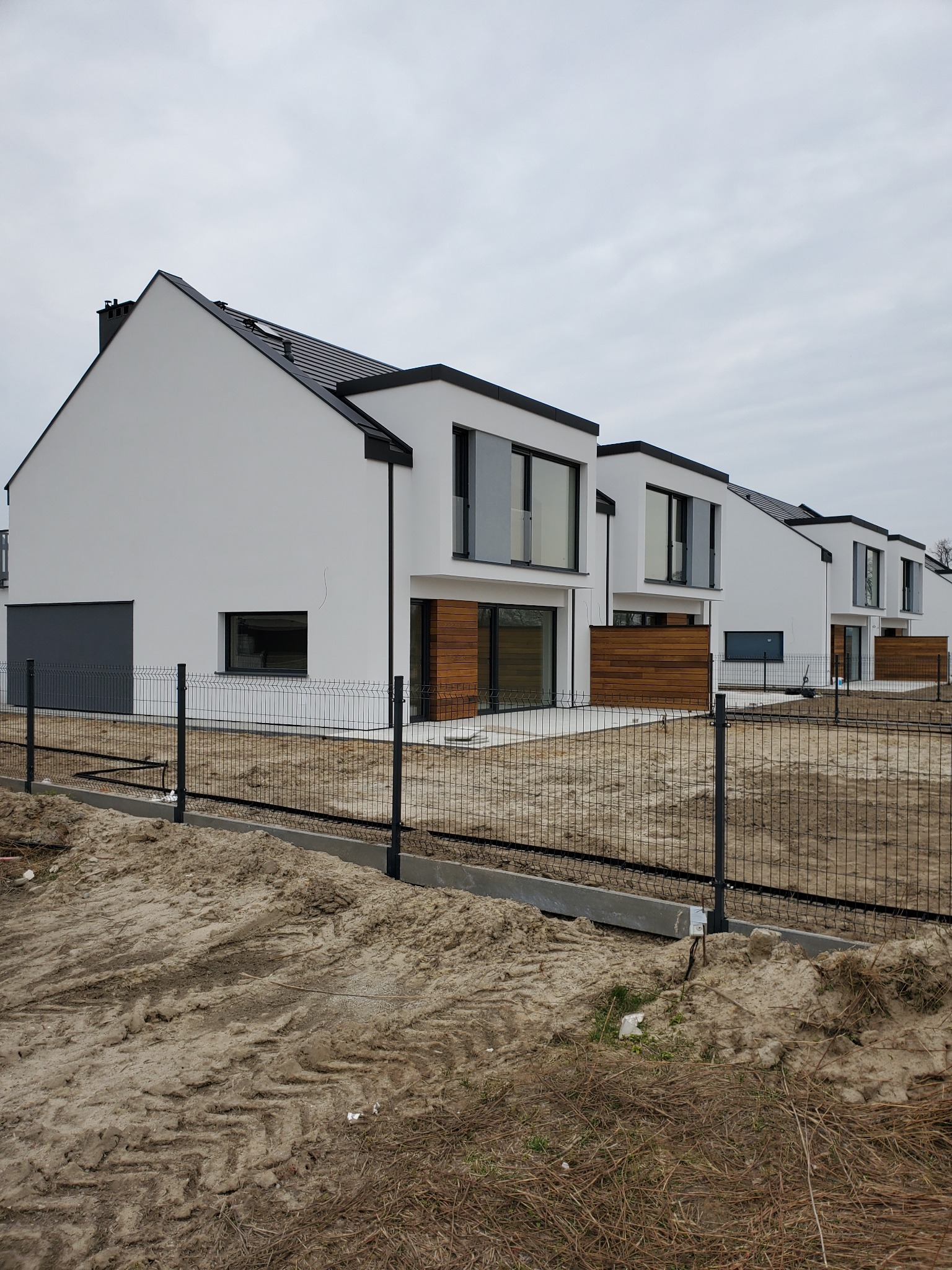 Rząd nowoczesnych, białych domów jednorodzinnych z płaskimi dachami i drewnianymi elementami elewacji, ogrodzonych czarną siatką na tle pochmurnego nieba.