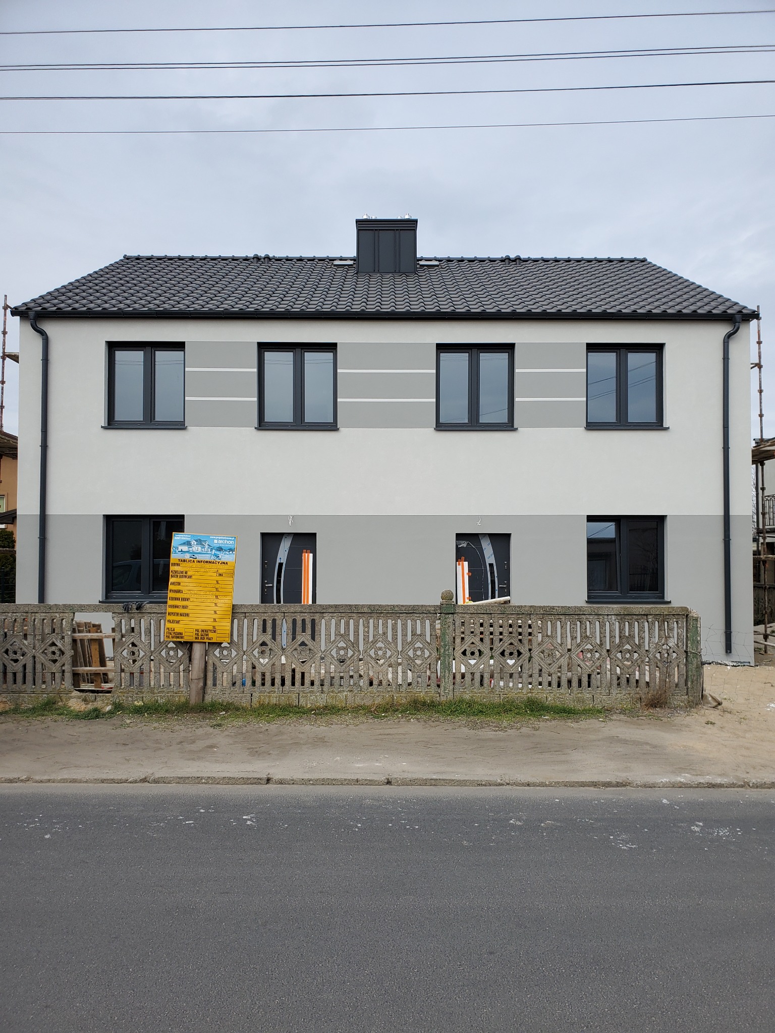 Dwukondygnacyjny dom z szarą elewacją i ciemnymi oknami, widoczny za betonowym płotem, z tablicą informacyjną budowy po lewej stronie.