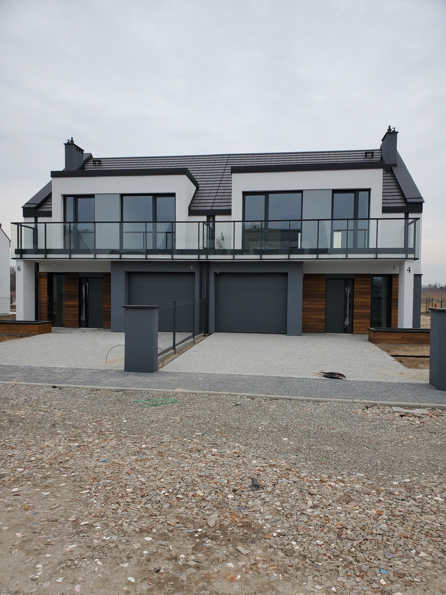 Nowoczesny bliźniak z białymi elewacjami, szarą stolarką okienną i balkonami z balustradami ze szkła, widok na niedokończony podjazd z gruzem.