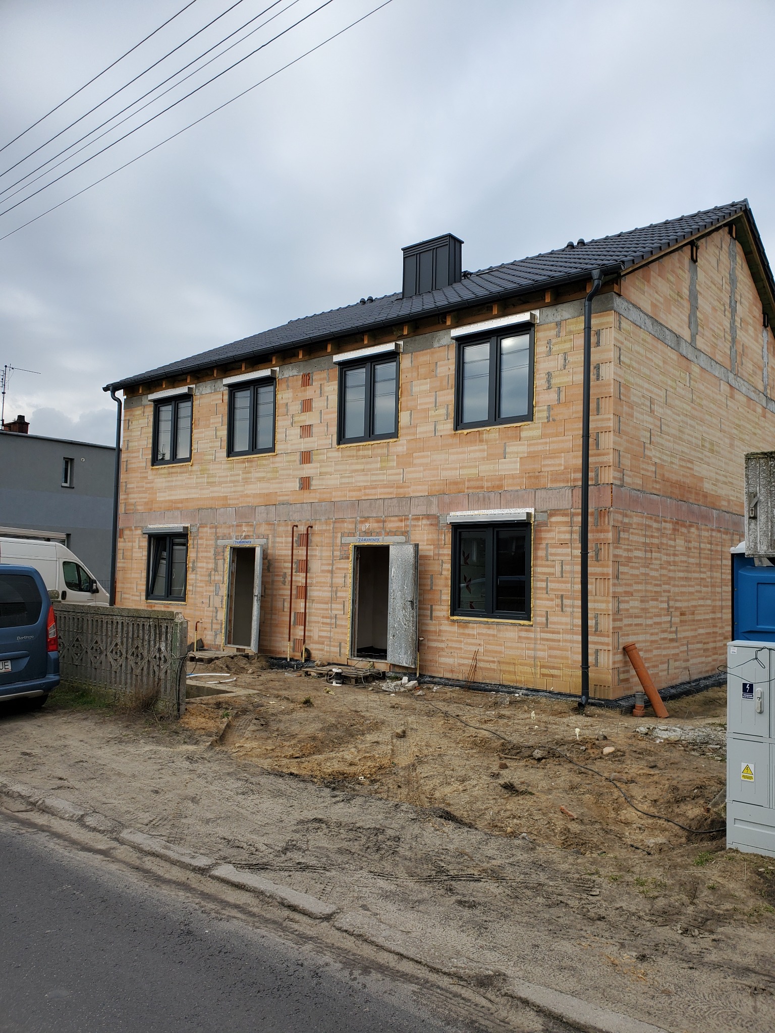 Surowy stan dwupiętrowego budynku z pustaków ceramicznych, z zamontowanymi czarnymi oknami i roletami zewnętrznymi, widoczne niezabezpieczone otwory drzwiowe i przygotowany teren wokół budynku...