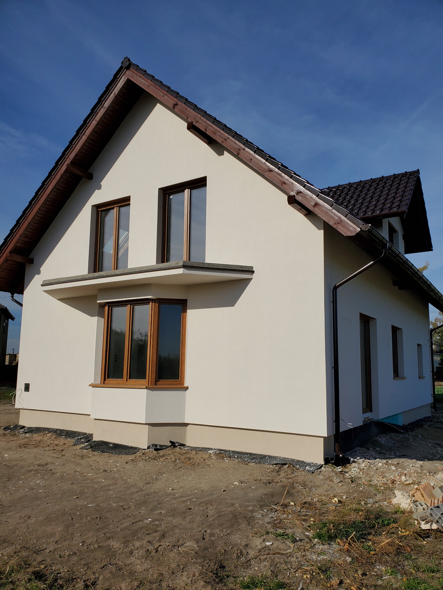 Nowoczesny dom jednorodzinny z jasną elewacją, brązowymi oknami i dachem, widok z zewnątrz na tle błękitnego nieba, teren wokół domu częściowo uporządkowany.