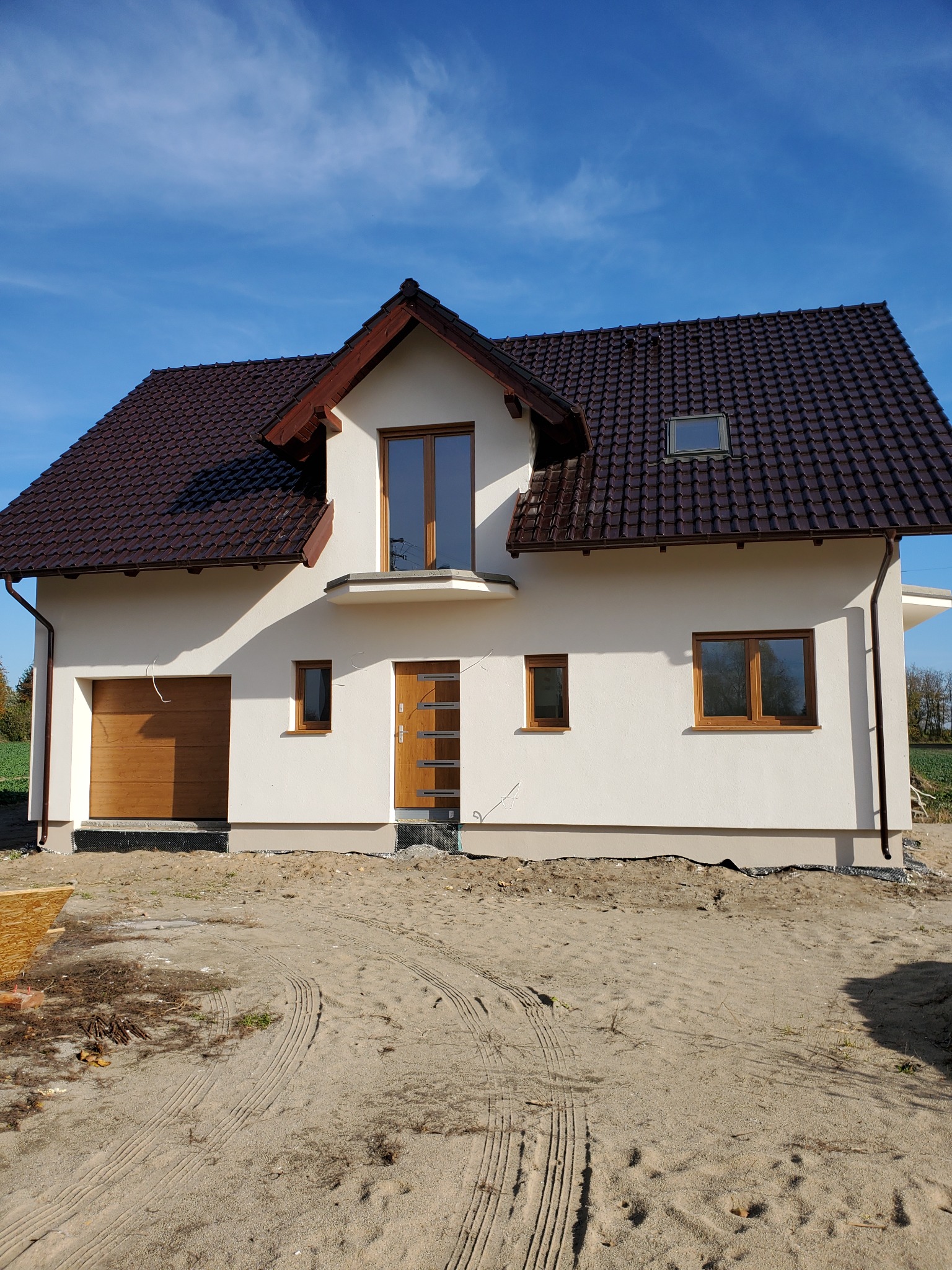 Nowoczesny dom jednorodzinny z brązowym dachem, jasną elewacją i drewnianymi oknami, widok z zewnątrz na tle błękitnego nieba, teren wokół domu w trakcie prac wykończeniowych.