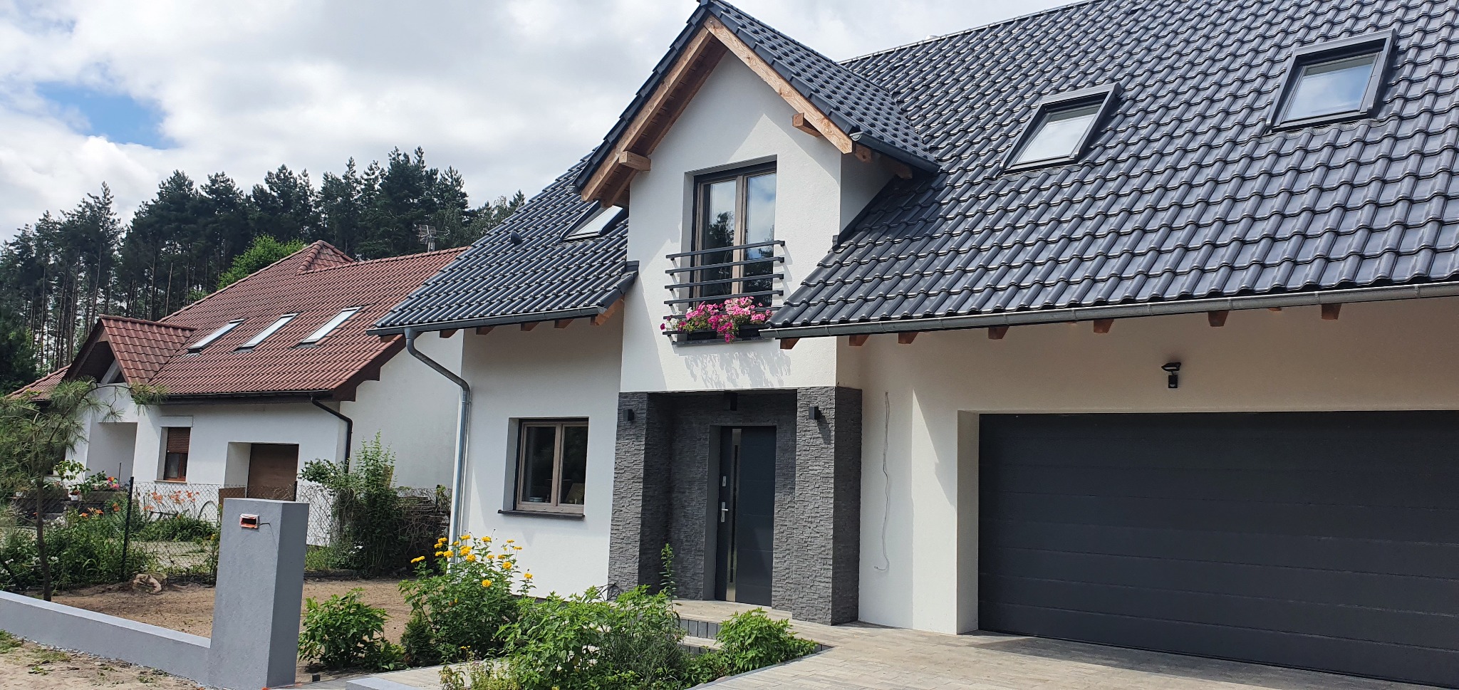 Nowoczesny dom jednorodzinny z białymi ścianami, ciemnoszarą elewacją imitującą kamień przy wejściu i garażu, balkonem z kwiatami i ciemną dachówką, na tle zielonego lasu i błękitnego nieba...