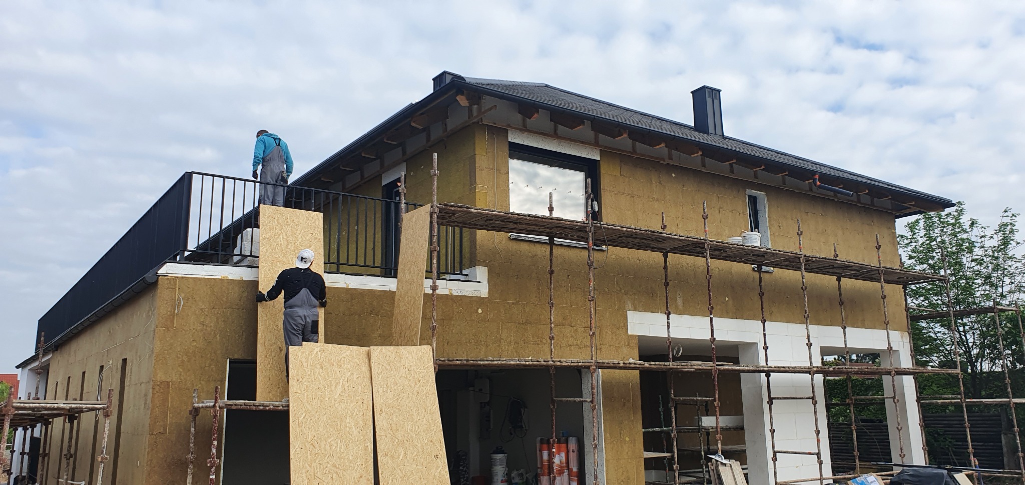 Dom w trakcie ocieplania: ściany pokryte wełną mineralną, rusztowania, pracownicy w kombinezonach i maskach montują płyty OSB.