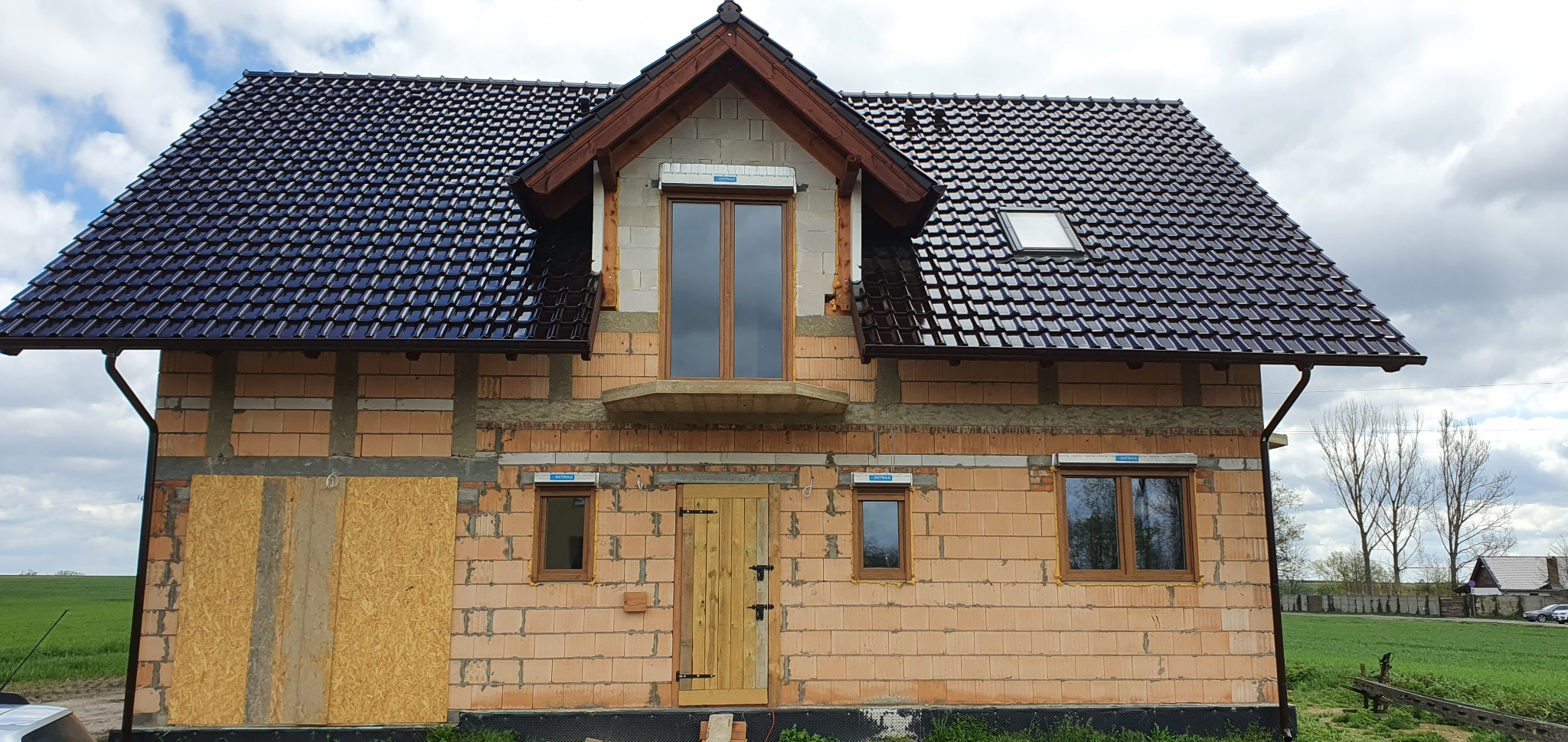 Niedokończony budynek jednorodzinny z czerwonej cegły, z zamontowanymi oknami z brązowymi ramami, drewnianymi drzwiami wejściowymi i czarną dachówką, jedna ze ścian zabita płytami OSB.