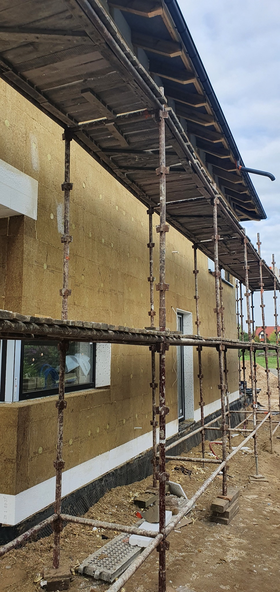 Dom w trakcie ocieplania, widoczne rusztowanie, izolacja termiczna na ścianach i fundamentach zabezpieczonych folią kubełkową.