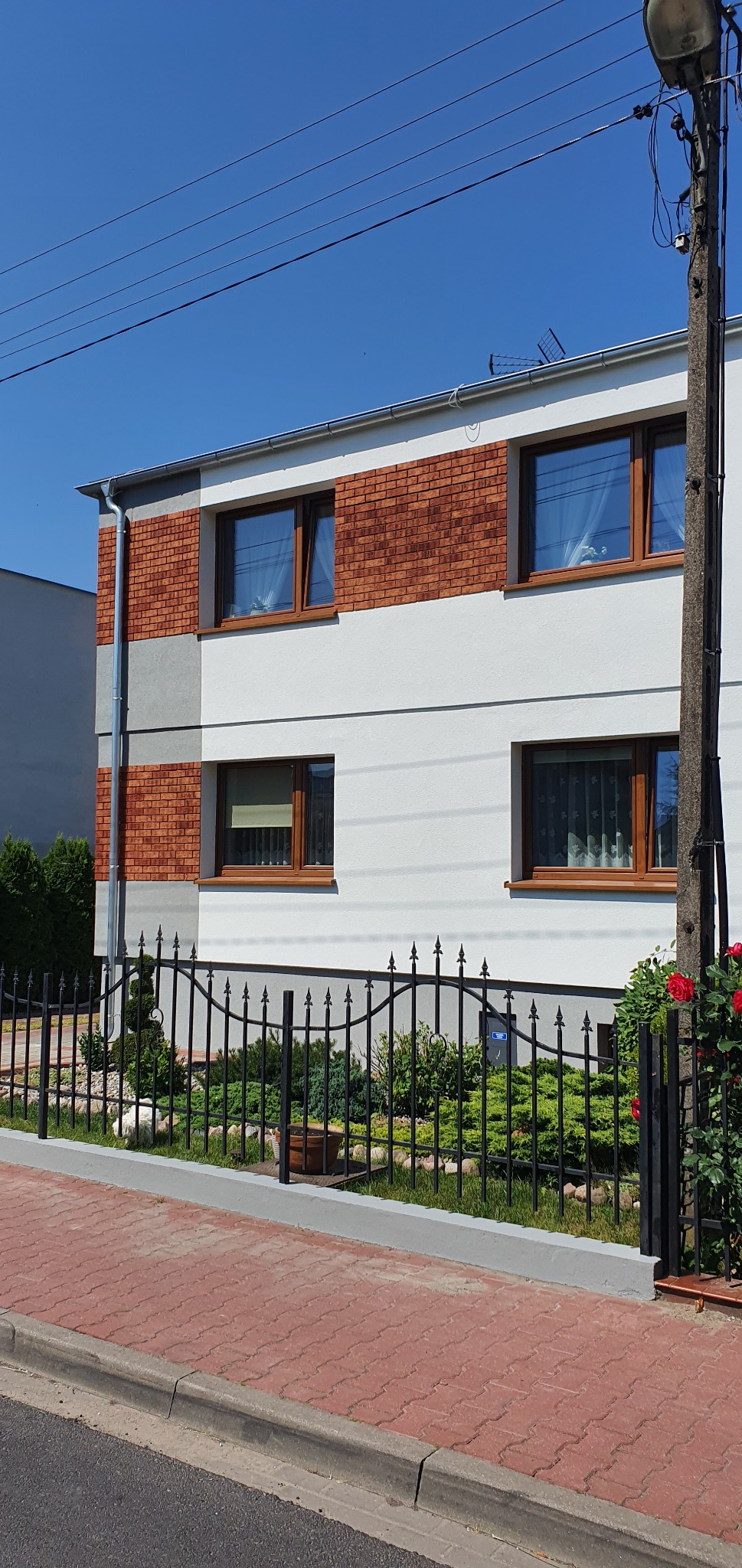 Dwukondygnacyjny dom z białą elewacją, fragmentami obłożonymi czerwoną cegłą klinkierową, widoczny zza czarnego, kutego ogrodzenia.