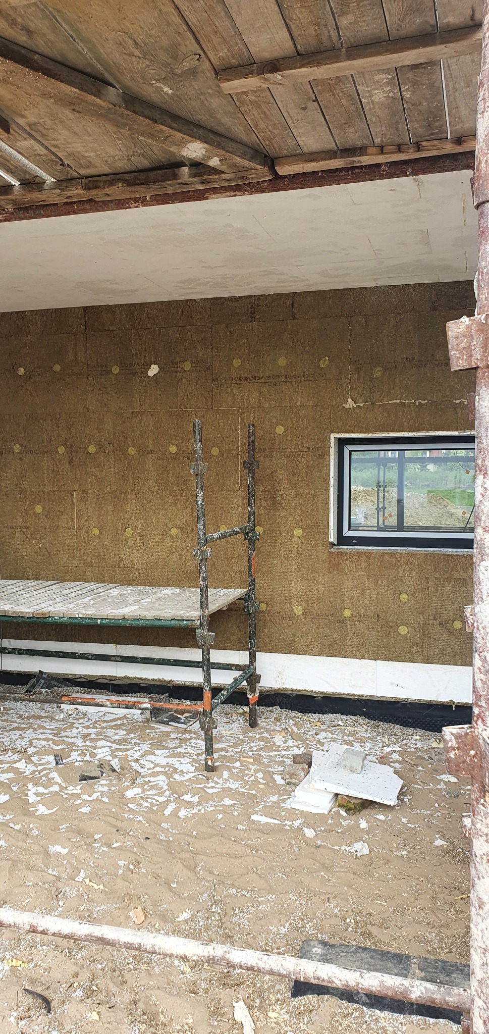 Ocieplanie elewacji wełną mineralną, widoczne rusztowanie, okno w czarnej ramie i fragment izolacji fundamentów przy ziemi.