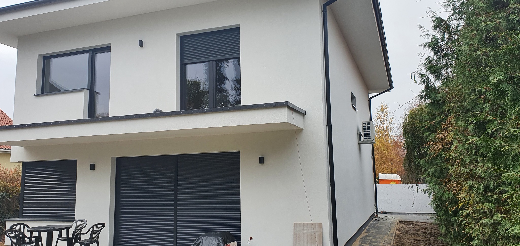 Dwukondygnacyjny dom z jasną, gładką elewacją, czarnymi ramami okiennymi i roletami, widok z boku na tle zieleni.