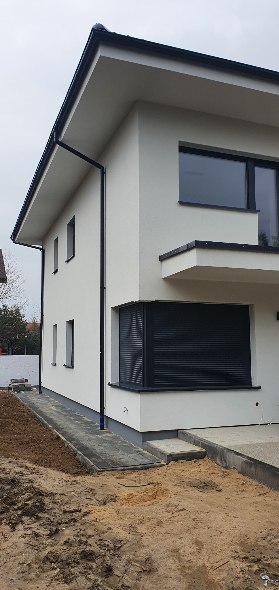 Nowoczesny dom z jasną elewacją, czarnymi ramami okiennymi i roletami zewnętrznymi, widoczne fragmenty chodnika i przygotowany teren wokół budynku.