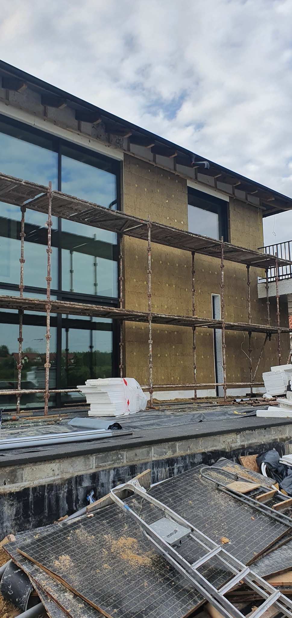 Dom w trakcie ocieplania, widoczne rusztowania, warstwa izolacyjna na ścianach, okna z czarnymi ramami oraz stos płyt izolacyjnych na dachu.