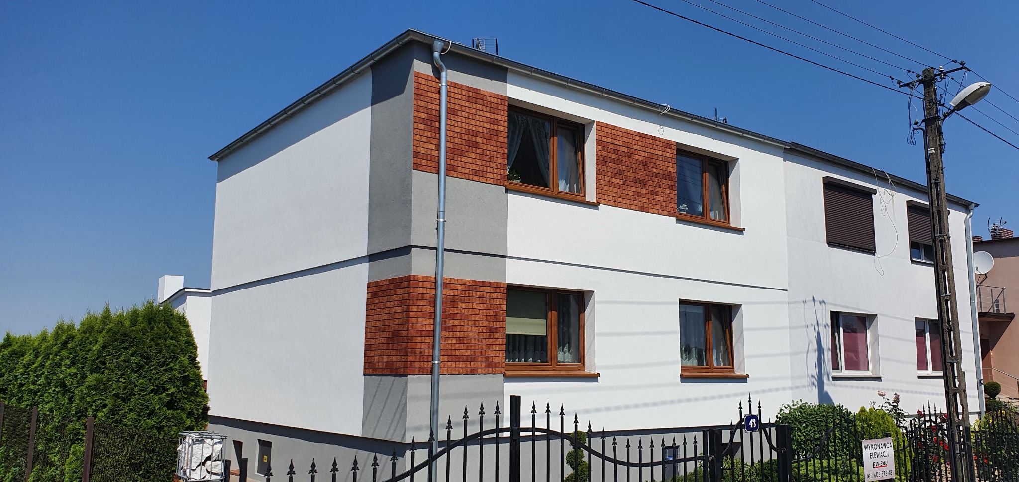 Dwupiętrowy dom z białą elewacją, ceglanymi akcentami i szarymi elementami dekoracyjnymi, widok z zewnątrz w słoneczny dzień.