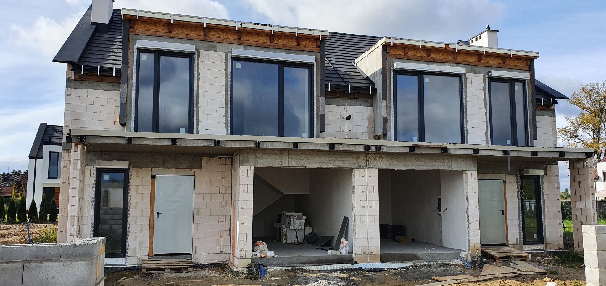 Niedokończony dom jednorodzinny z białych bloczków, widoczne okna z roletami zewnętrznymi, brak tynku, elementy konstrukcyjne dachu w trakcie montażu, otwarte garaże na parterze.