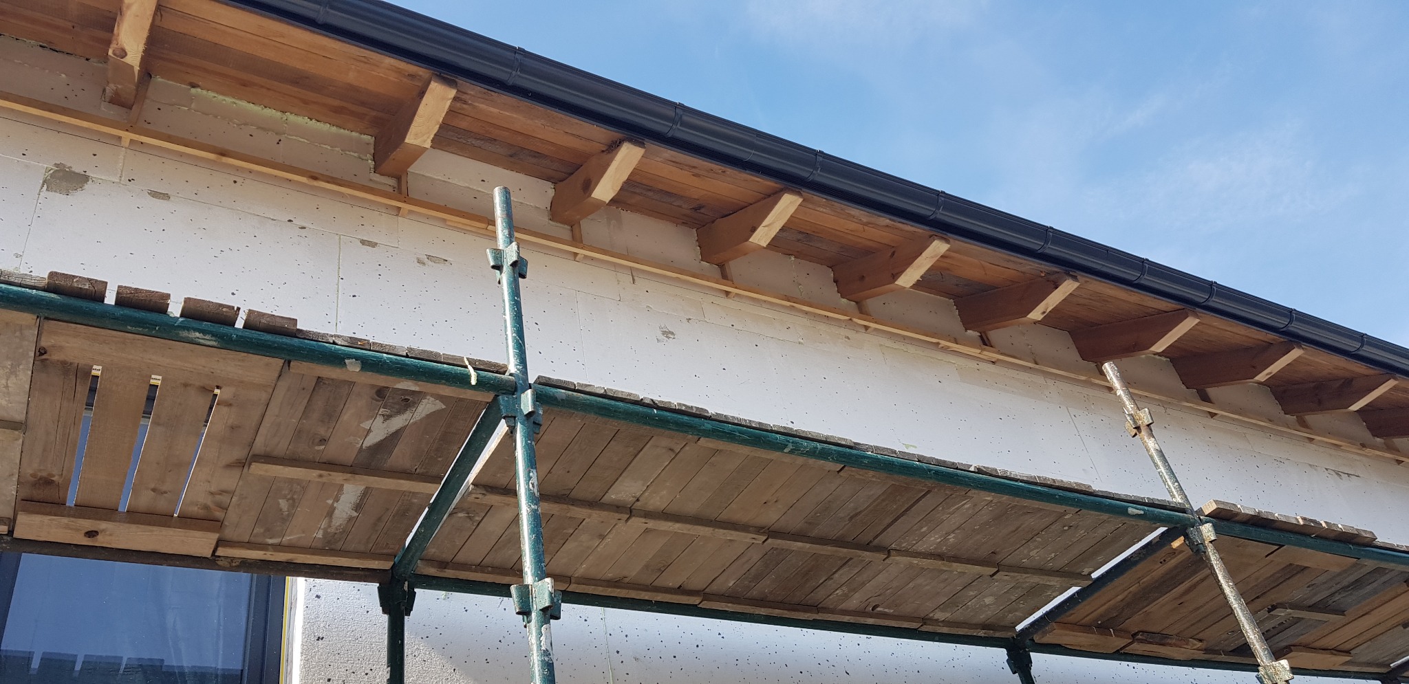 Ściana budynku w trakcie prac, widoczne rusztowanie, styropianowe płyty izolacyjne oraz drewniane elementy konstrukcyjne dachu i rynna.