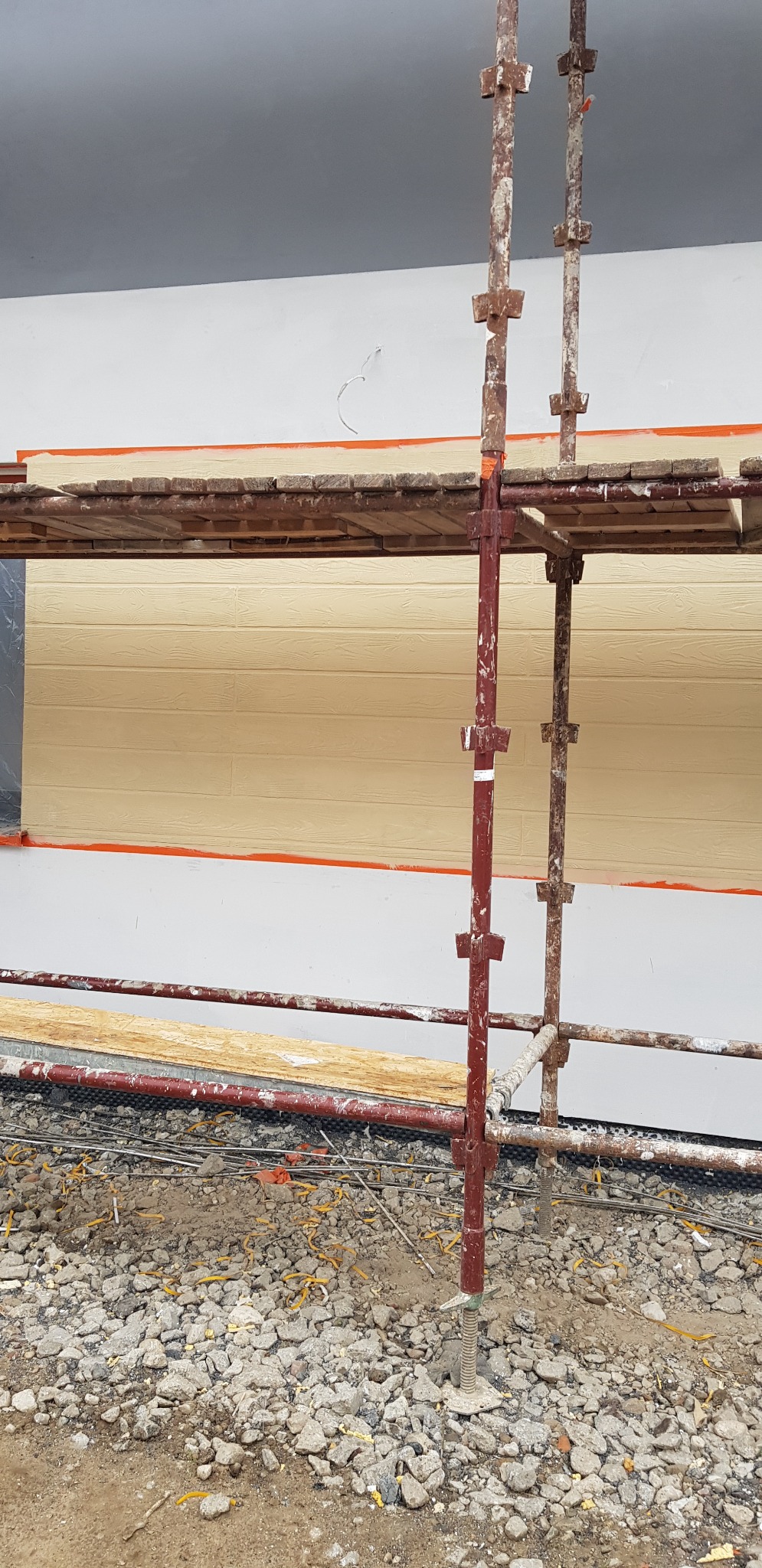 Fragment elewacji z widocznym rusztowaniem podczas prac wykończeniowych, dolna część ściany w kolorze kremowym z imitacją drewna, górna część biała, zabezpieczona pomarańczową taśmą malarską.