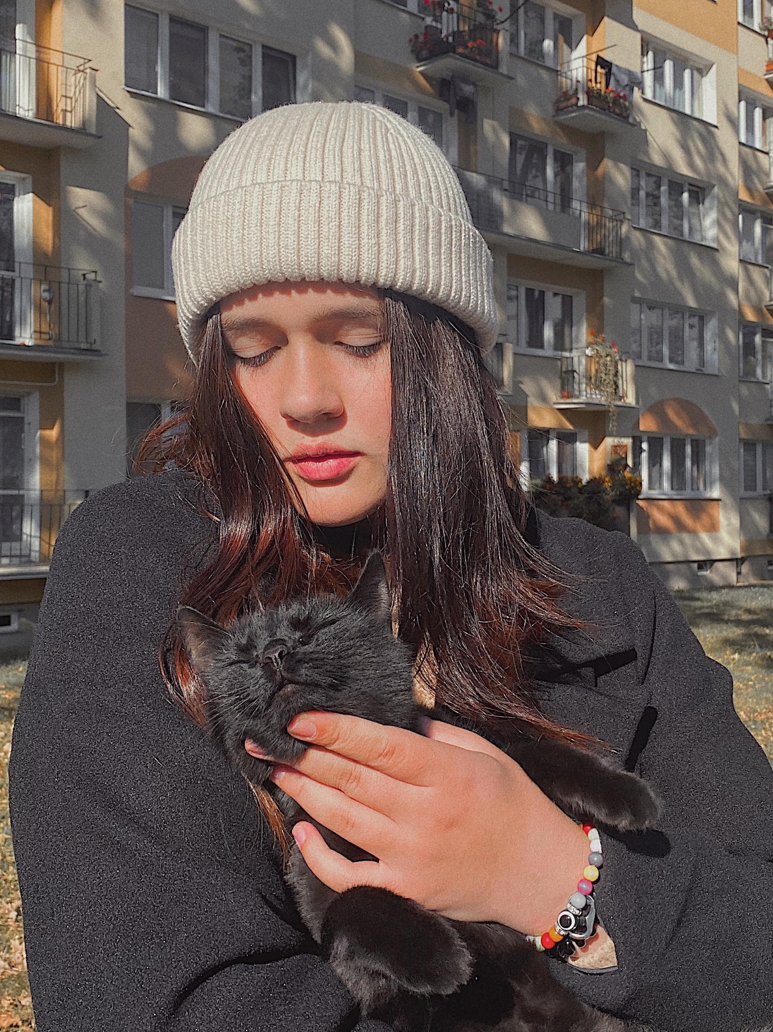 Młoda kobieta w białej czapce trzyma na rękach czarnego kota na tle bloku mieszkalnego w słoneczny dzień.