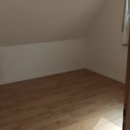 Poddasze po remoncie z jasnymi panelami podłogowymi, świeżo pomalowanymi białymi ścianami i oknem z żaluzjami, widoczne zza otwartych drzwi.