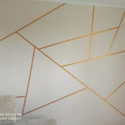 Biała ściana z geometrycznym wzorem wykonanym złotą farbą, widoczny fragment łóżka z szarą pościelą w lewym dolnym rogu.