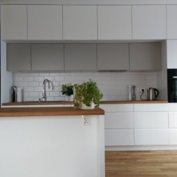 Lewkowicz Meble Kuchnie&Szafy - Nowoczesna kuchnia z białymi szafkami, drewnianym blatem i czarną tablicą kredową po lewej stronie, z rośliną w doniczce na wyspie.