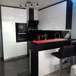 Lewkowicz Meble Kuchnie&Szafy - Nowoczesna kuchnia z białymi, lakierowanymi frontami, czarnym blatem i barkiem z czerwonym akcentem, oświetlona listwą LED pod szafkami wiszącymi, z dwoma czarnymi hokerami przy barku.