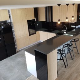 Lewkowicz Meble Kuchnie&Szafy - Nowoczesna kuchnia z jasnymi drewnianymi frontami, czarnym blatem i sprzętem AGD, oświetlona trzema wiszącymi lampami z geometrycznymi kloszami