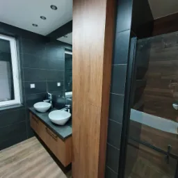 Nowoczesna łazienka z dwoma białymi umywalkami nablatowymi, szafką pod umywalkę z drewnianym frontem, wysoką drewnianą szafą i kabiną prysznicową z czarnymi profilami.