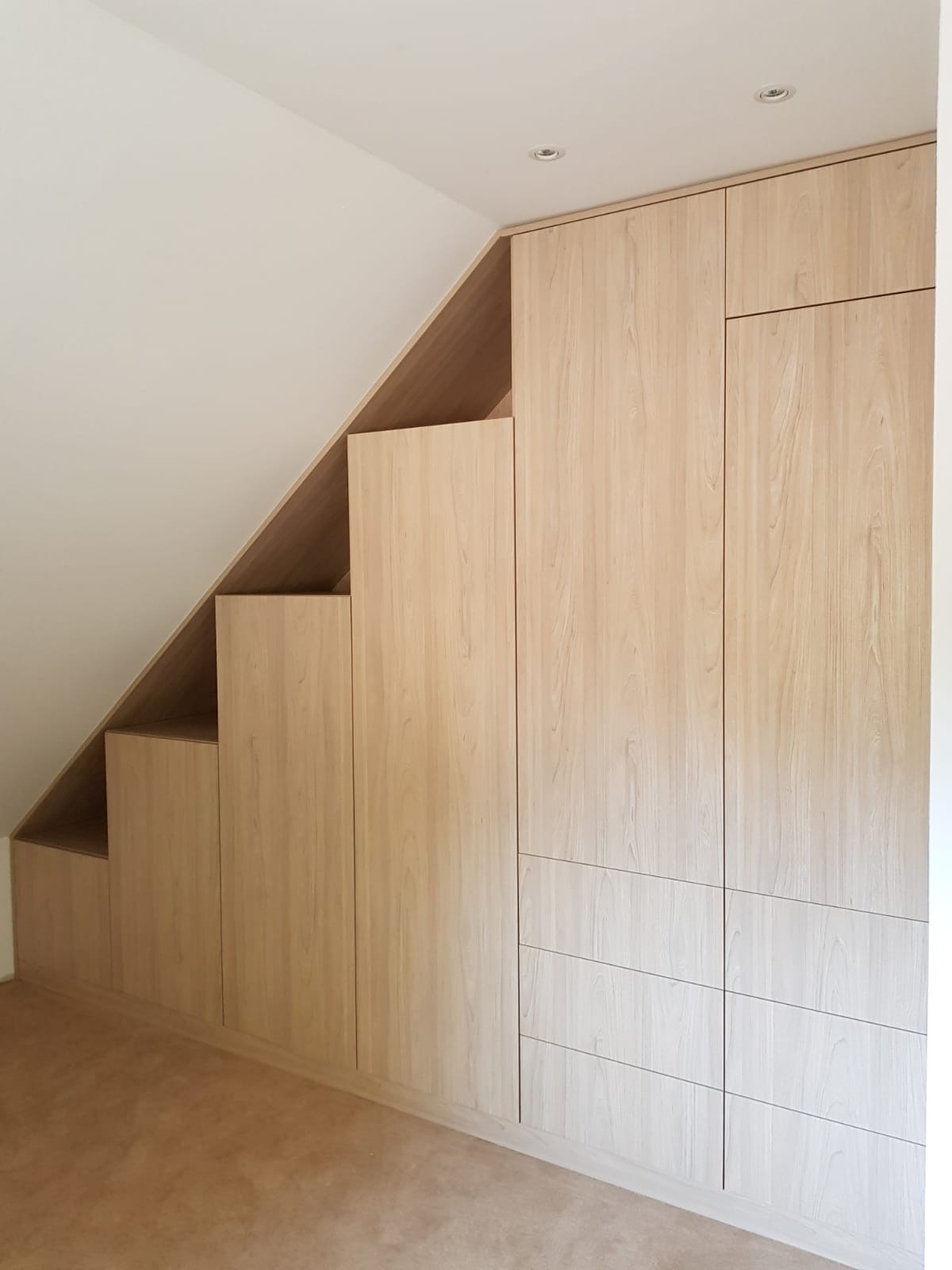 Szafa wnękowa z jasnego drewna, zabudowa pod skosem dachu, zintegrowana z szufladami, oświetlenie punktowe na suficie, minimalistyczny design.