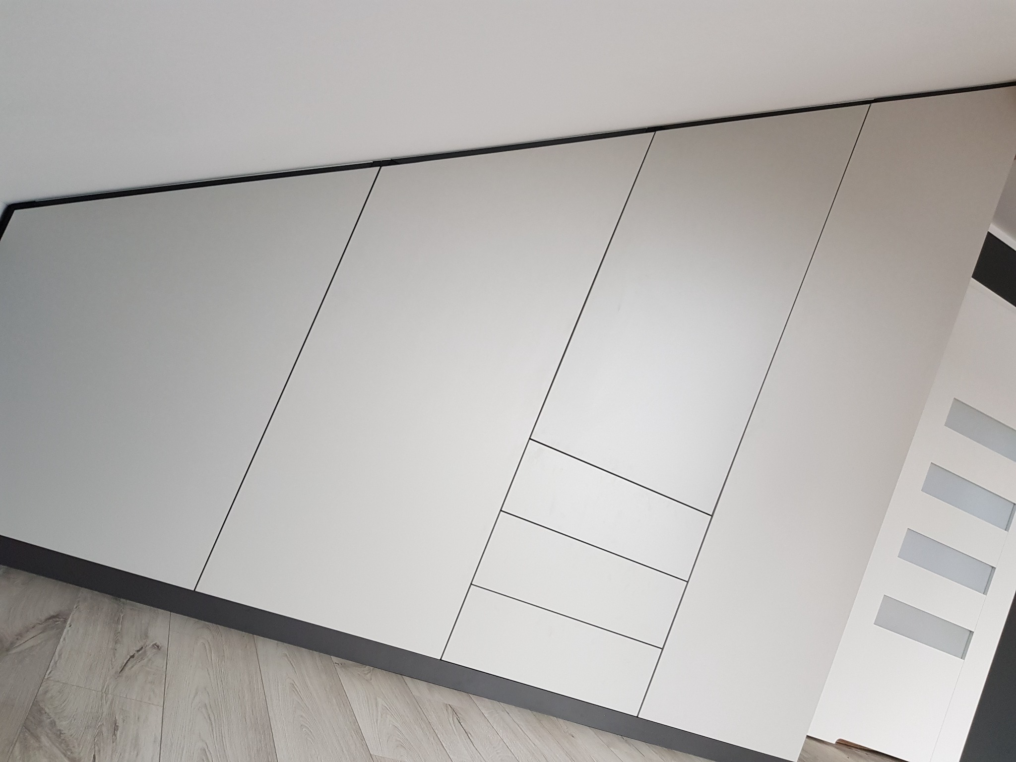 Biała szafa wnękowa z ukośnymi drzwiami i trzema szufladami, czarne obramowanie, widok od dołu, jasna podłoga z paneli.