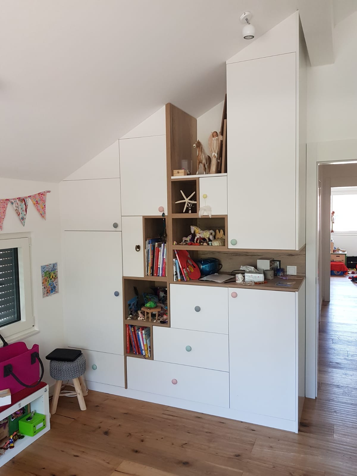 Biała zabudowa meblowa na poddaszu ze schodkowymi półkami i kolorowymi gałkami, wypełniona książkami i zabawkami dziecięcymi.