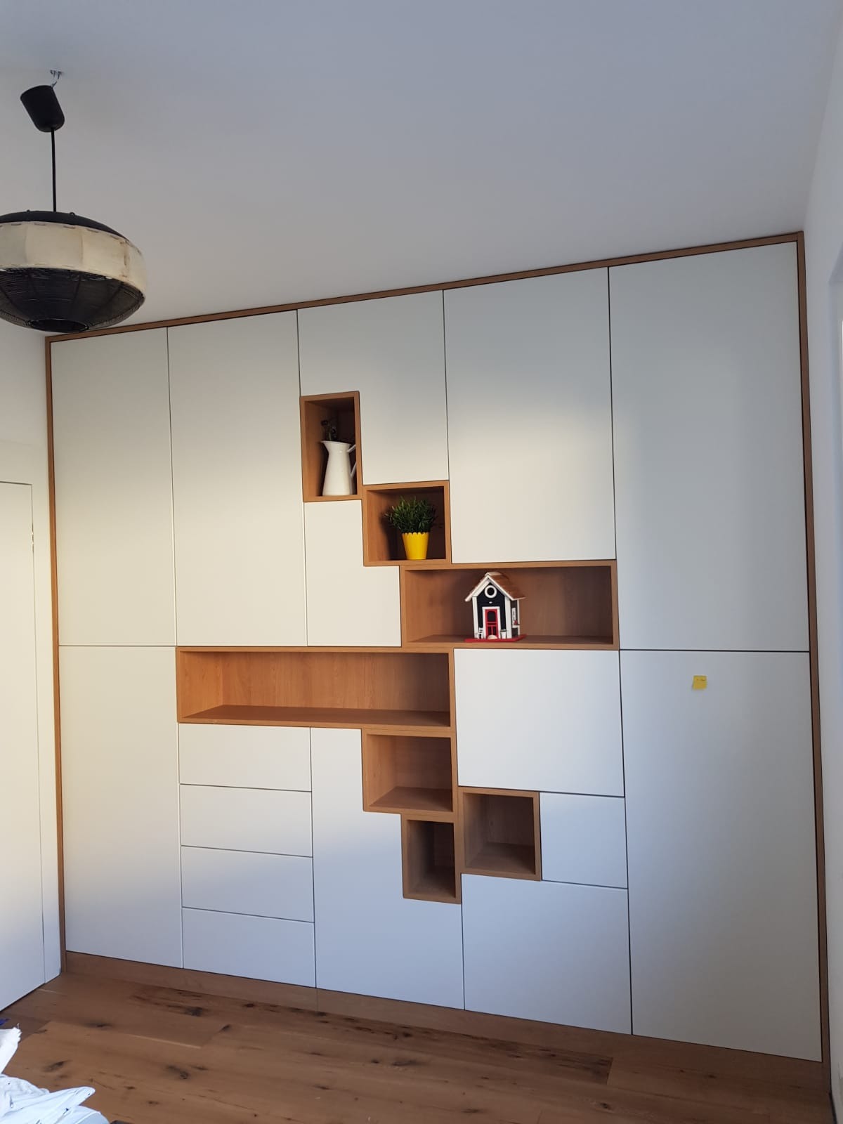 Biała szafa wnękowa z drewnianymi wstawkami, asymetrycznymi półkami, dekoracjami: dzbankiem, doniczką i domkiem, na drewnianej podłodze.